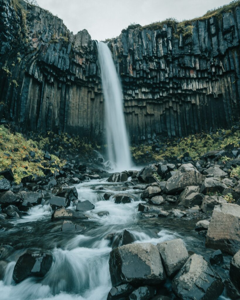 Majestic waterfall cascading over volcanic basalt cliffs with river stones and lush greenery wallpaper