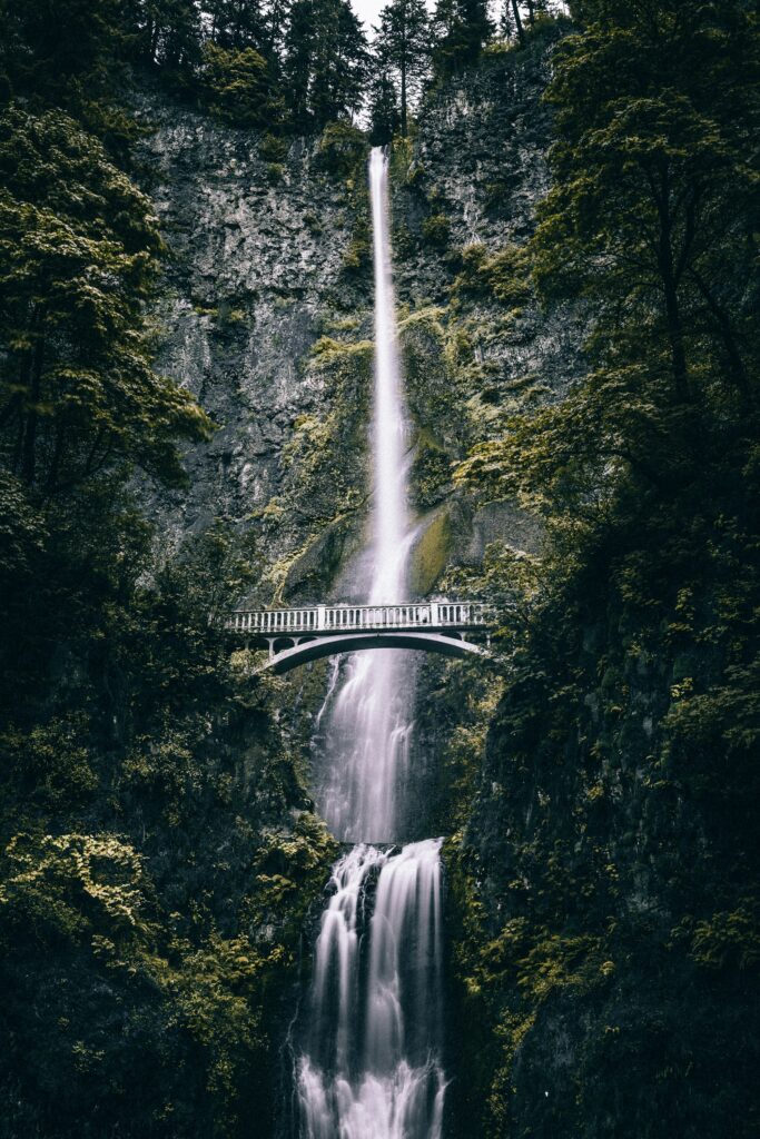Majestic waterfall flows under arched bridge surrounded by lush green cliffs in serene landscape Wallpaper