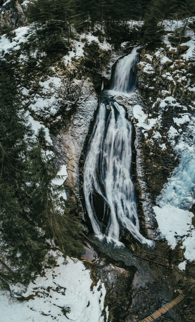 Aerial view of a winter waterfall flowing through snow-covered rocks in evergreen forest landscape Wallpaper