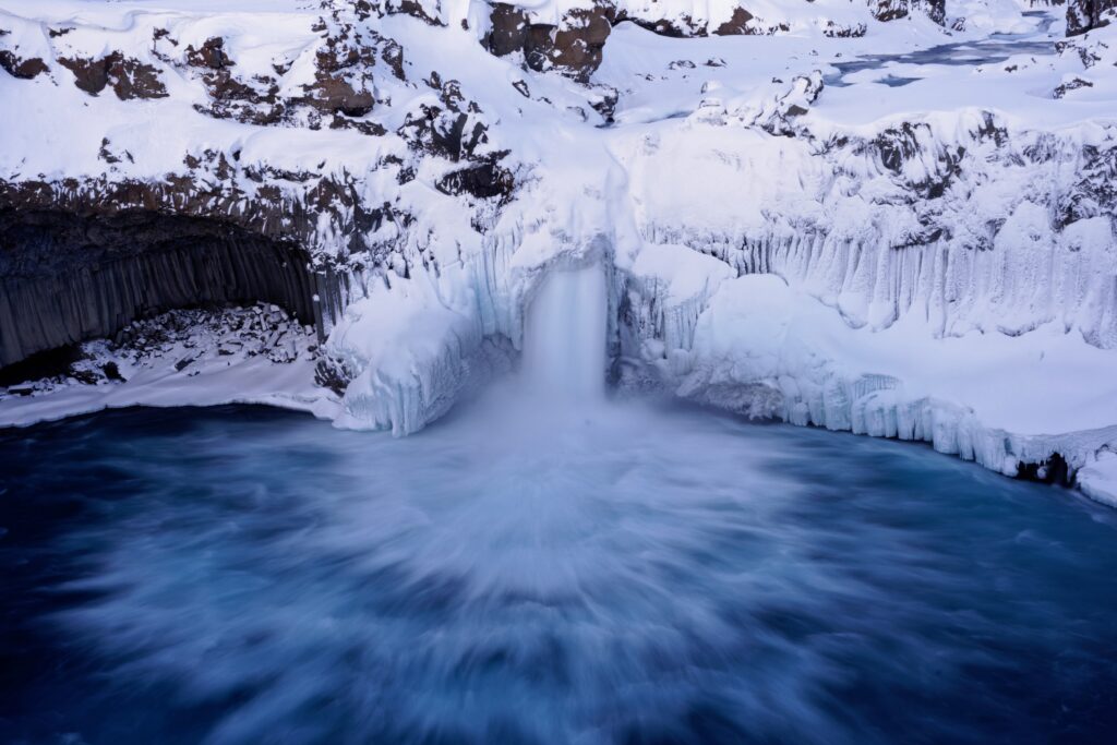 Majestic waterfall crashing into icy pool surrounded by snow-covered cliffs and blue icebergs Wallpaper