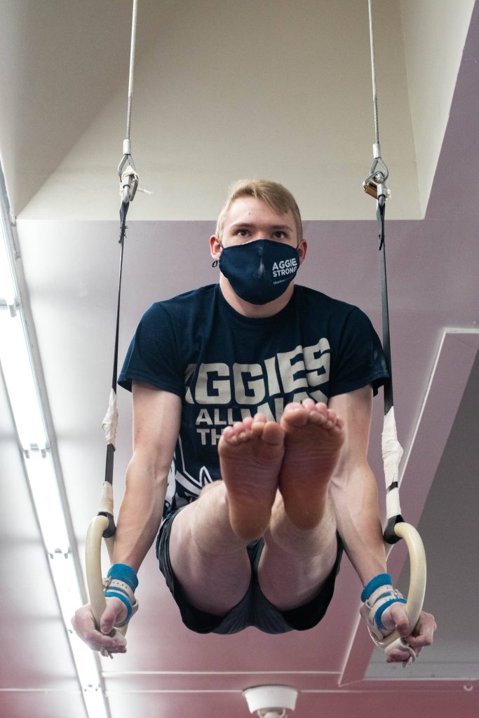 Male gymnast in Aggies shirt performing L-sit on rings showing core strength and discipline Wallpaper