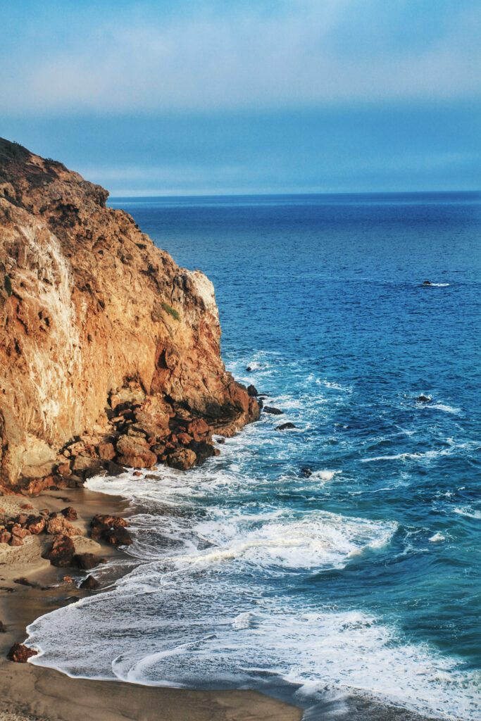 Coastal Encounter: Majestic Waves Crashing Against Malibu Cliffside in Sunlit Serenity Wallpaper