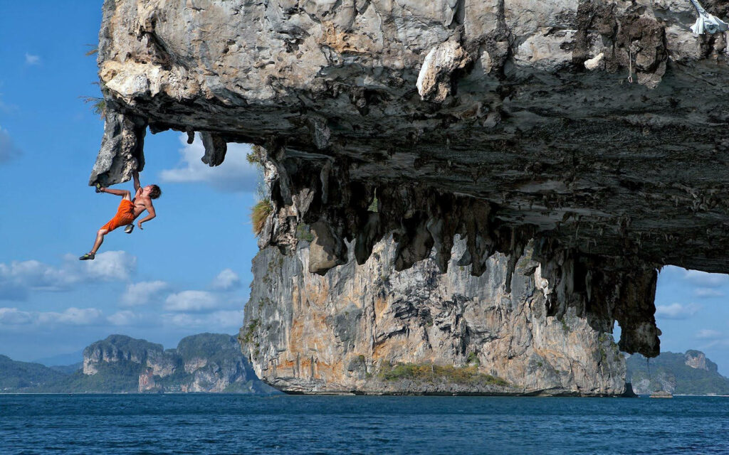 Man in bright orange shorts climbing rugged rock overhang above blue sea with limestone cliffs Wallpaper