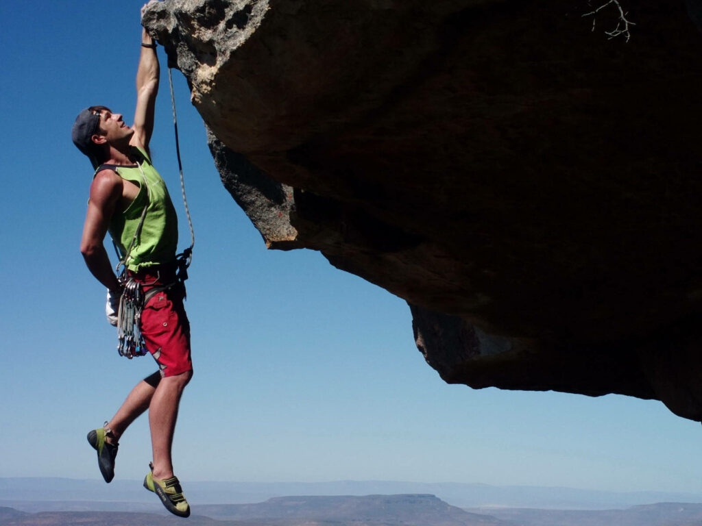 Man hanging one-handed from steep rock face showing climbing strength and skill against blue sky Wallpaper