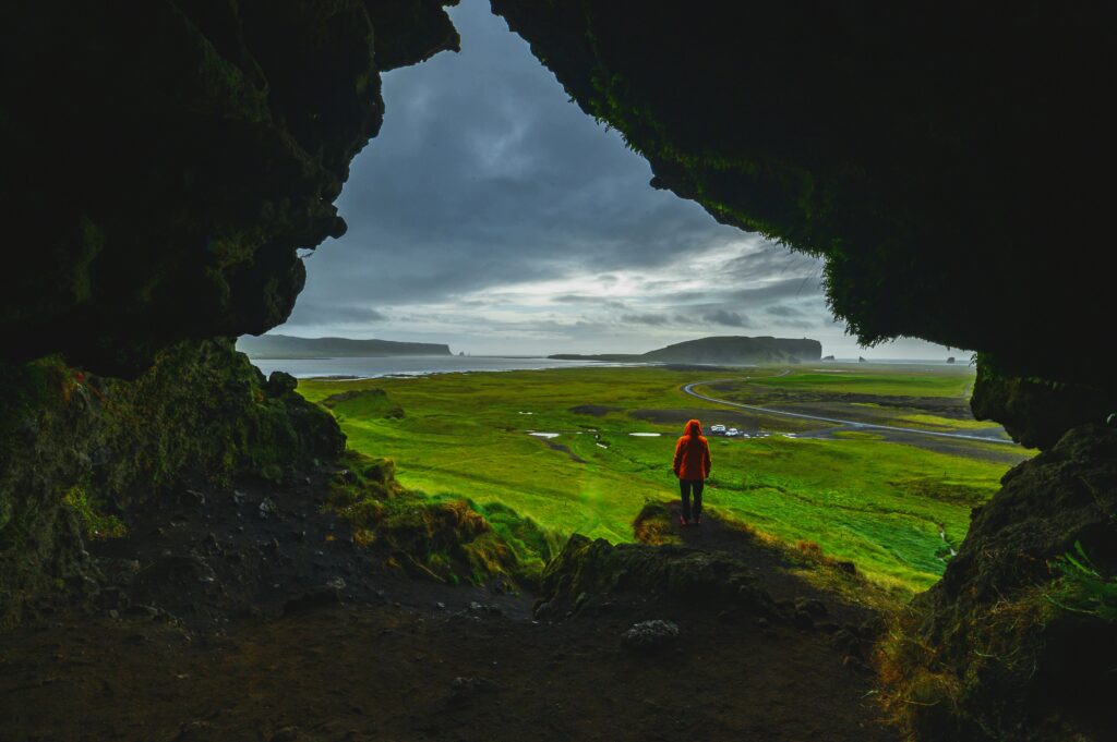 Man in orange jacket at dark cave entrance overlooking lush coastal landscape with winding roads Wallpaper