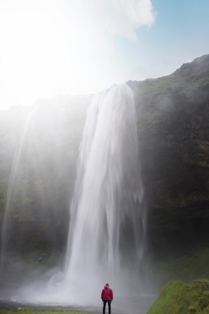 Man in red jacket views majestic waterfall with sunlight and mist over lush green landscape Wallpaper