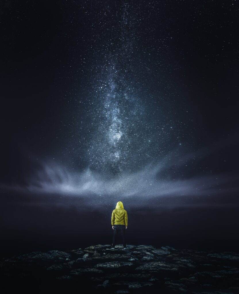 Man in yellow jacket on rocky terrain gazing at starry night sky with Milky Way above wallpaper