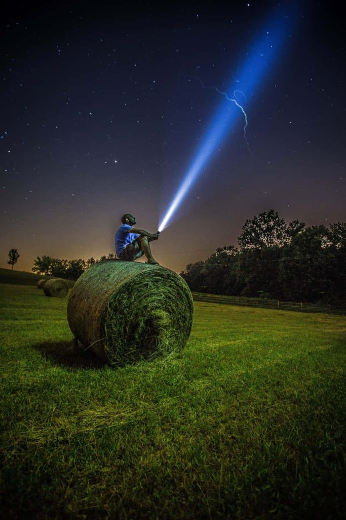 Man on hay bale shining flashlight into starry night sky rural field serene wallpaper background