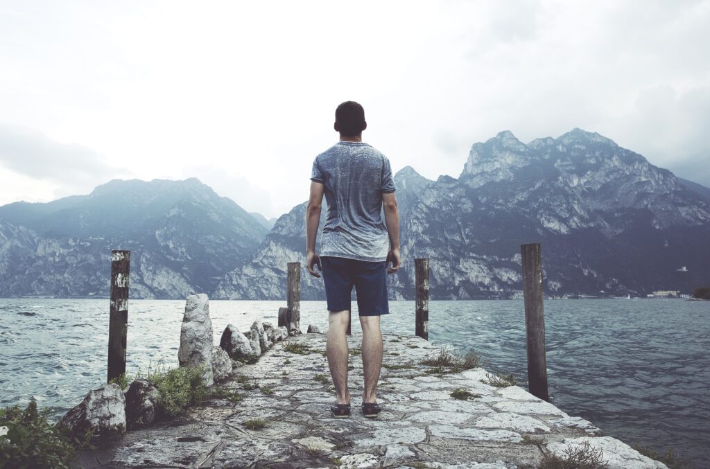 Man on rustic stone pier overlooking vast sea and towering mountains in tranquil blue tones Wallpaper