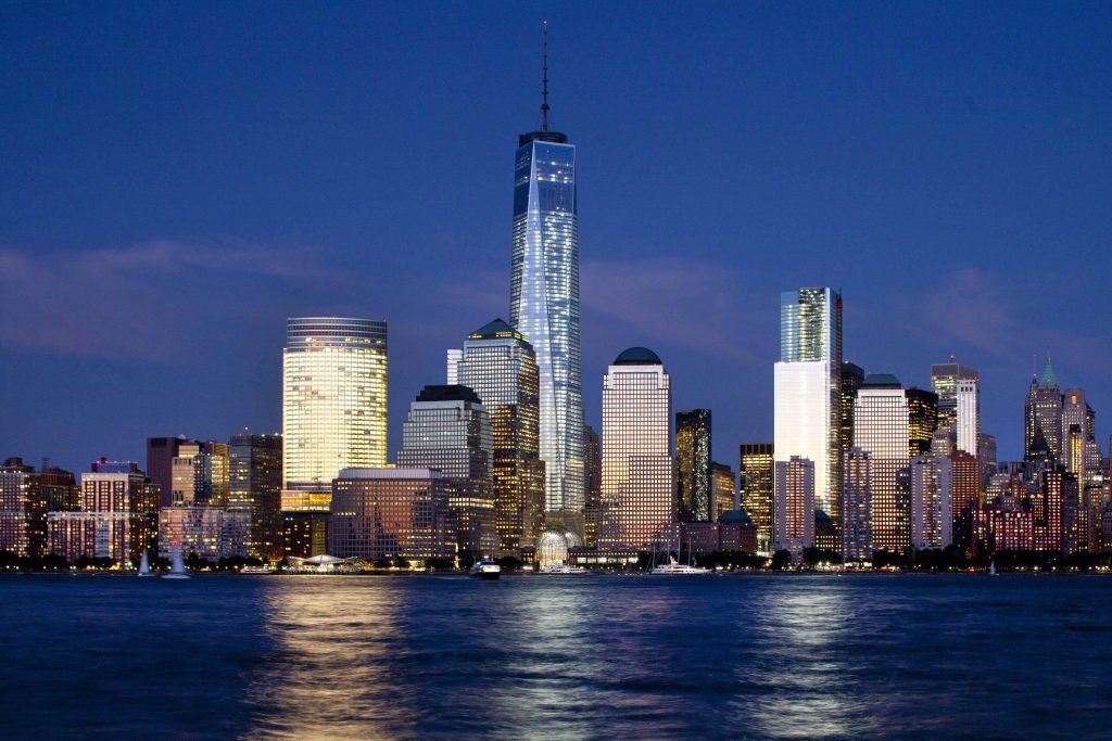 Manhattan skyline with illuminated One World Trade Center at twilight reflecting on calm water wallpaper