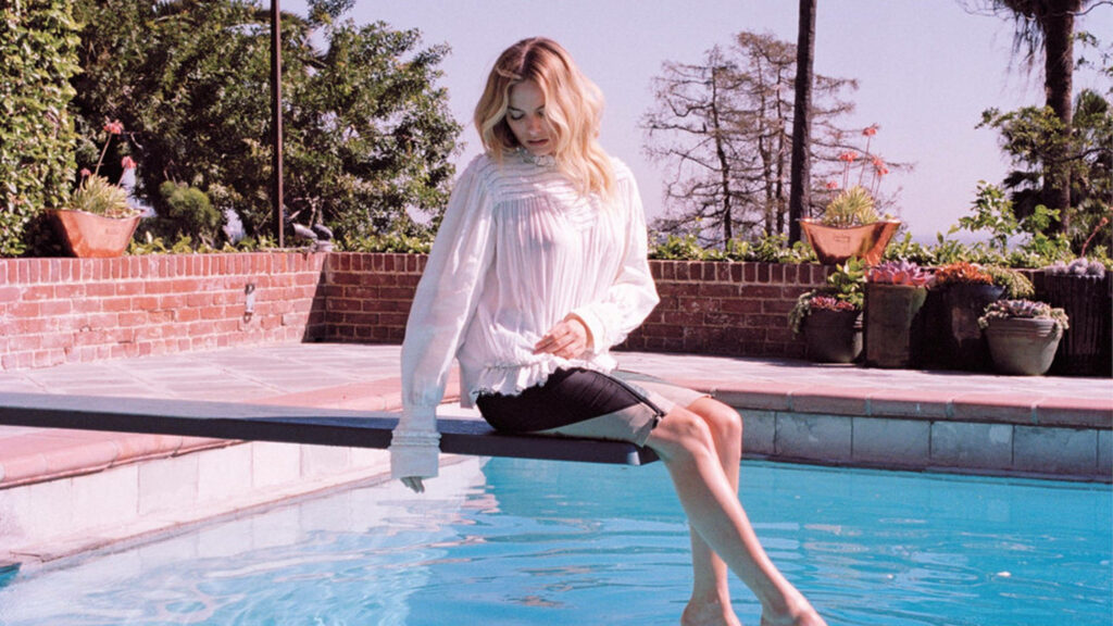 Margot Robbie sitting on a pool springboard dipping feet in water surrounded by greenery and sunlight Wallpaper