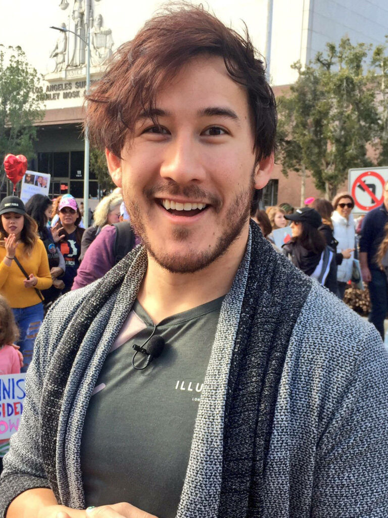 Markiplier smiling at outdoor fan convention with colorful signs lively crowd wallpaper background