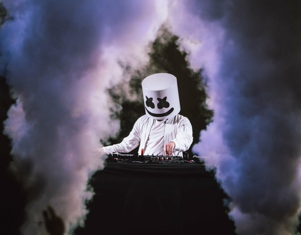 DJ Marshmello mixing on Pioneer decks with helmet on stage surrounded by smoke and lights Wallpaper