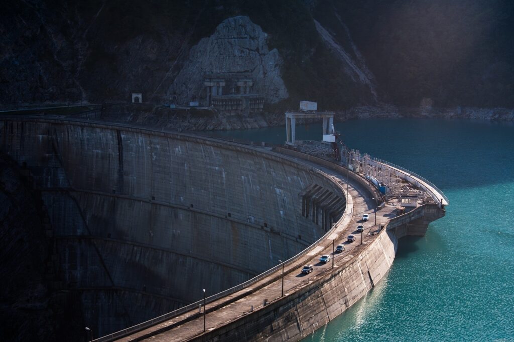 Massive curved concrete dam over turquoise river with rugged cliffs and vehicles on road at daylight Wallpaper