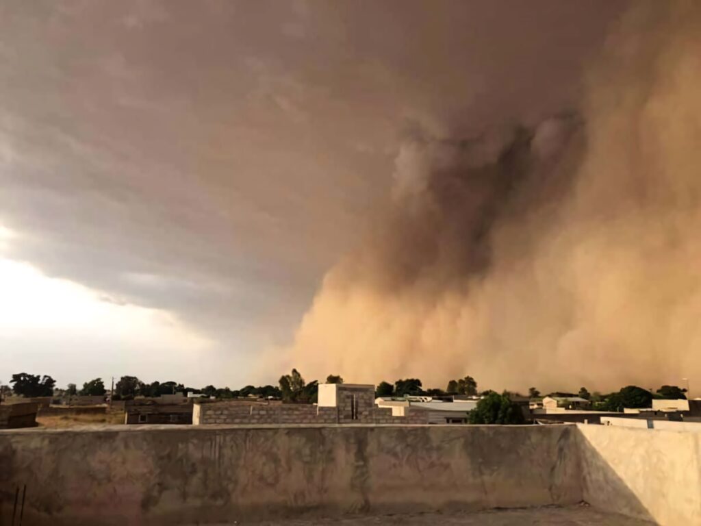 Massive dust storm looms over quiet village with sunlit buildings in vivid brown gray haze Wallpaper