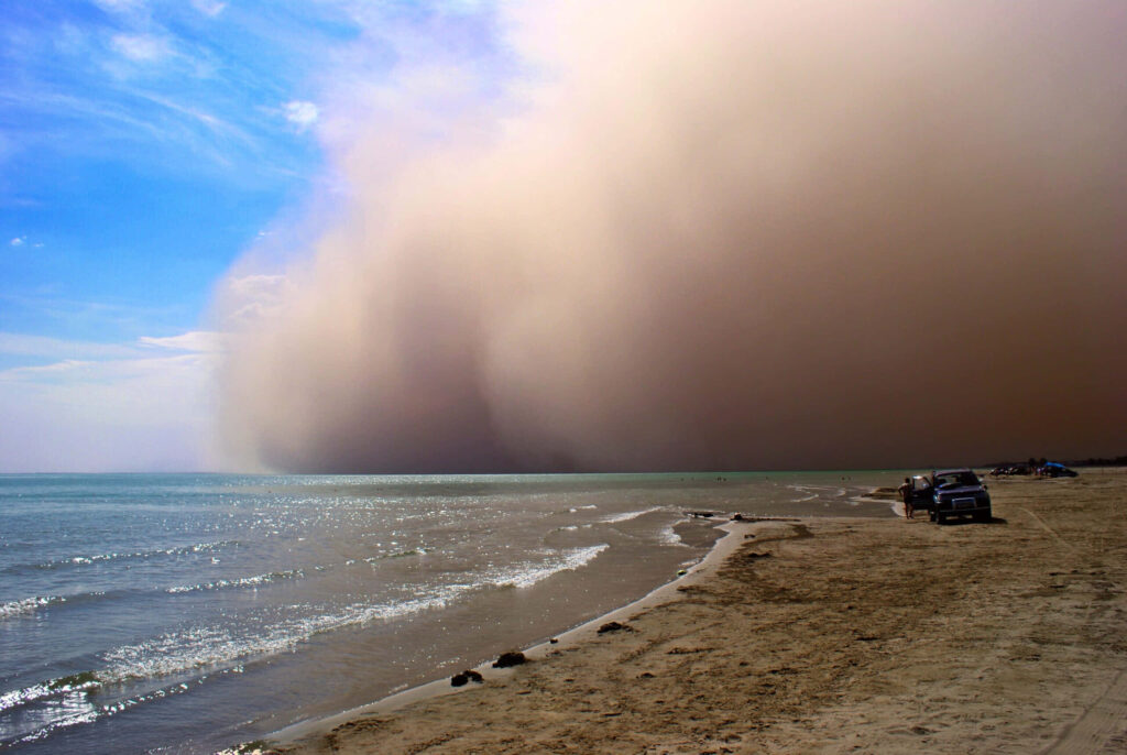 Massive dust storm approaches serene beach with turquoise water golden sand and blue sky backdrop Wallpaper