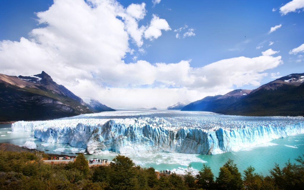 Massive glacier with icy blue blocks over turquoise lake surrounded by mountains and tourists Wallpaper