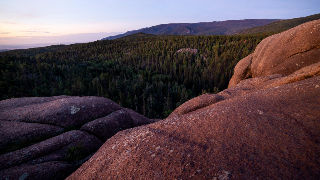 Massive reddish stones in foreground with dense pine forest and rolling hills under evening sky Wallpaper