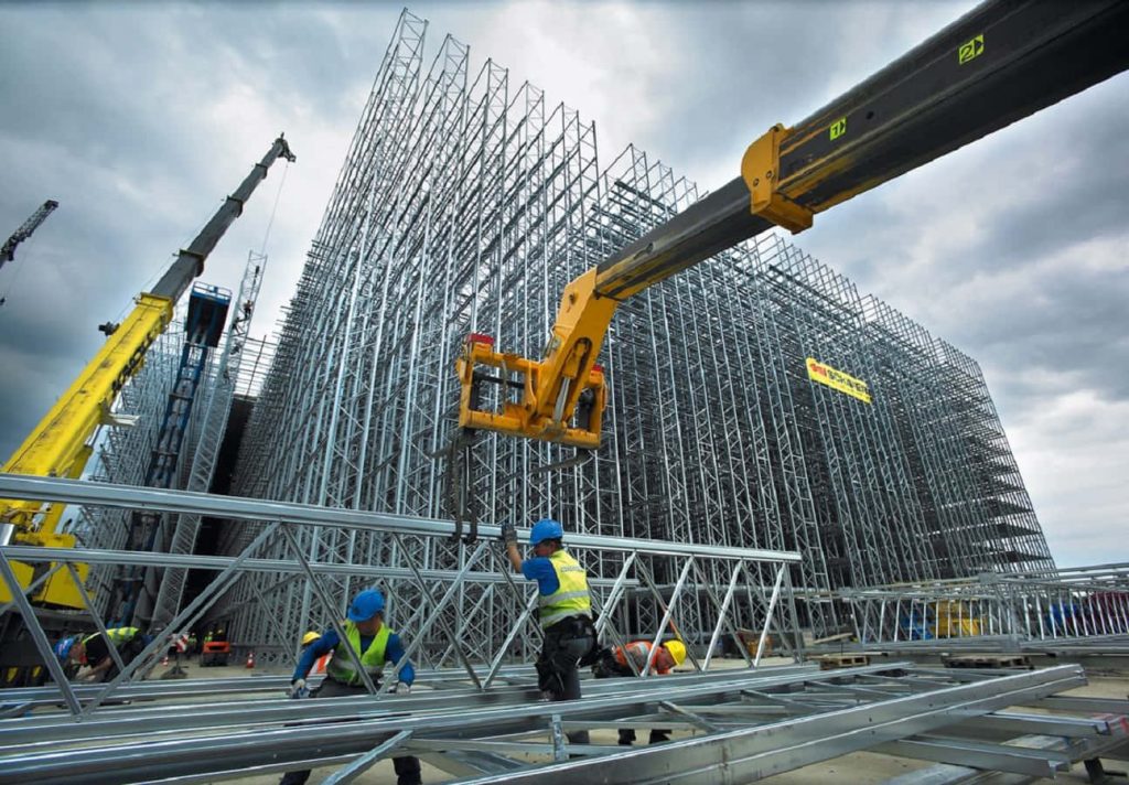 Workers assembling massive SSI Schäfer high-bay racking with yellow crane and steel framework Wallpaper