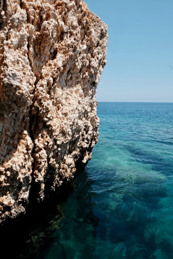 Massive textured coastal rock formation above clear turquoise sea with sunlight enhancing colors Wallpaper