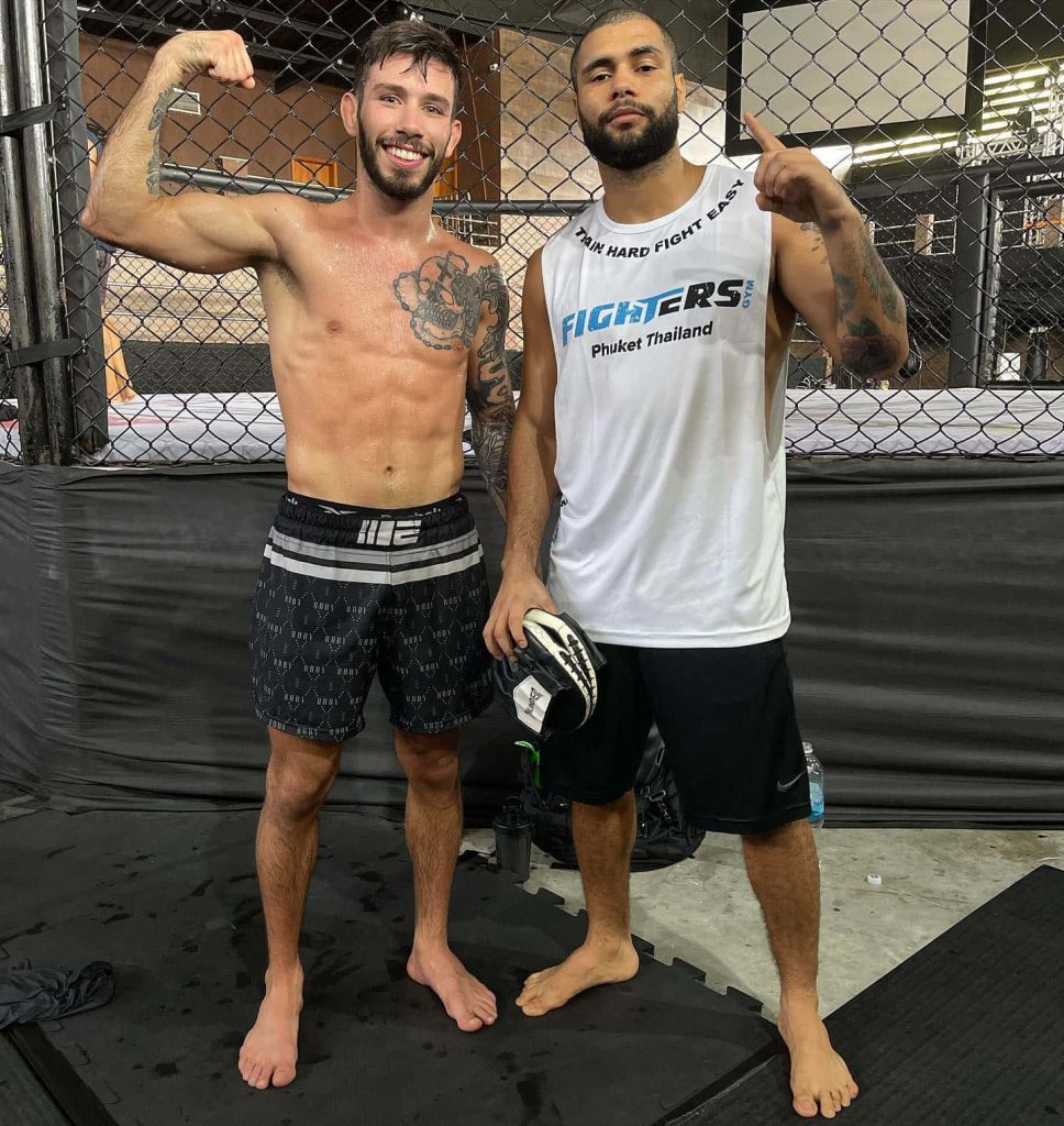 UFC fighter Matheus Nicolau flexes shirtless with training partner in MMA gym octagon background Wallpaper