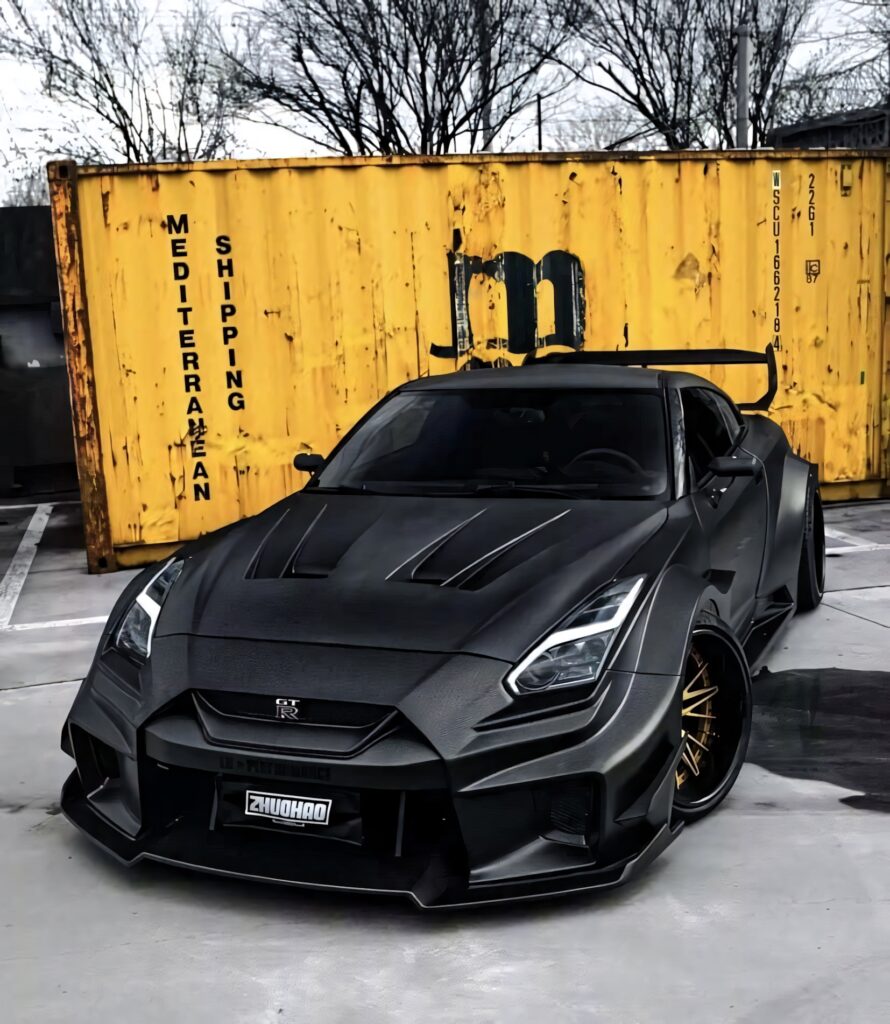 Matte black Nissan R35 GTR with aggressive body kit and gold wheels parked by yellow container Wallpaper