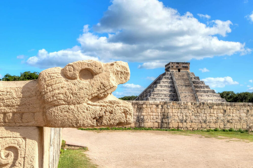 Maya snake head carving with Kukulkan pyramid under bright blue sky at Chichen Itza wallpaper