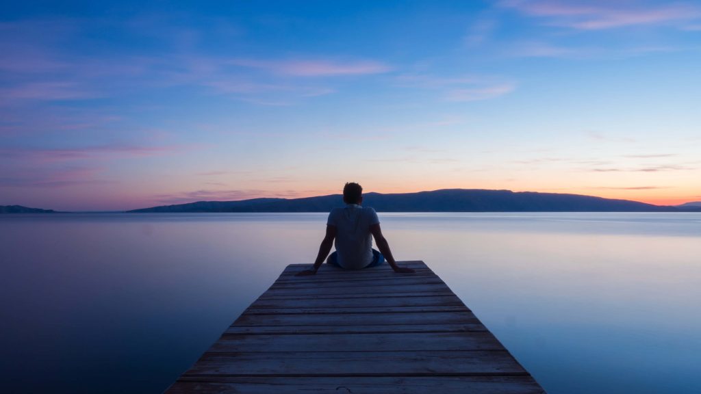 Lone sad man on wooden pier at sunset calm sea blue orange hues contemplative wallpaper background