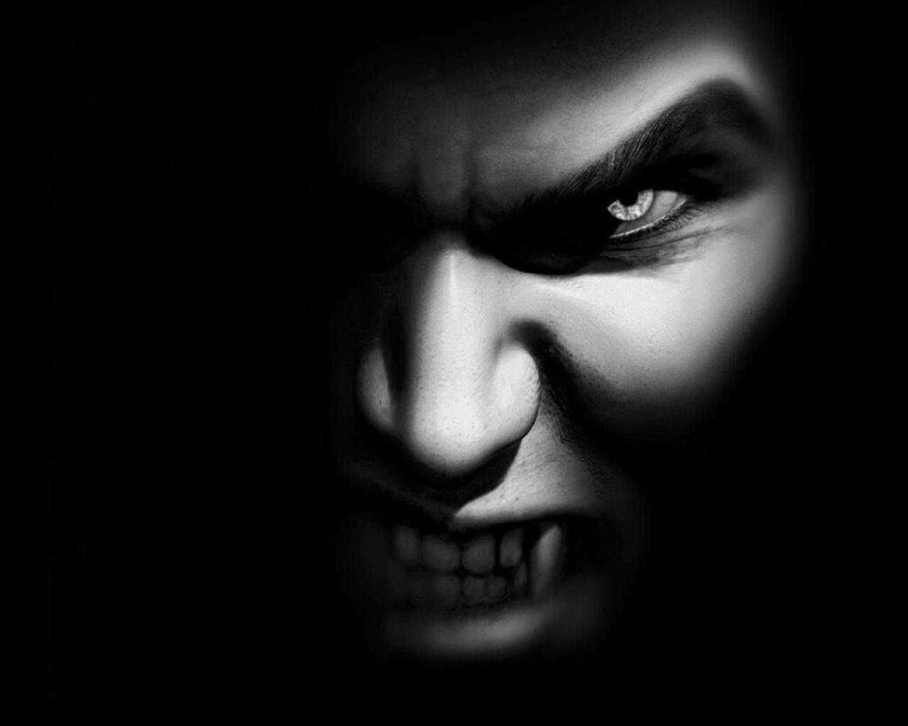 Menacing vampire face with sharp fangs and fierce eyes emerging from dark shadowy background Wallpaper