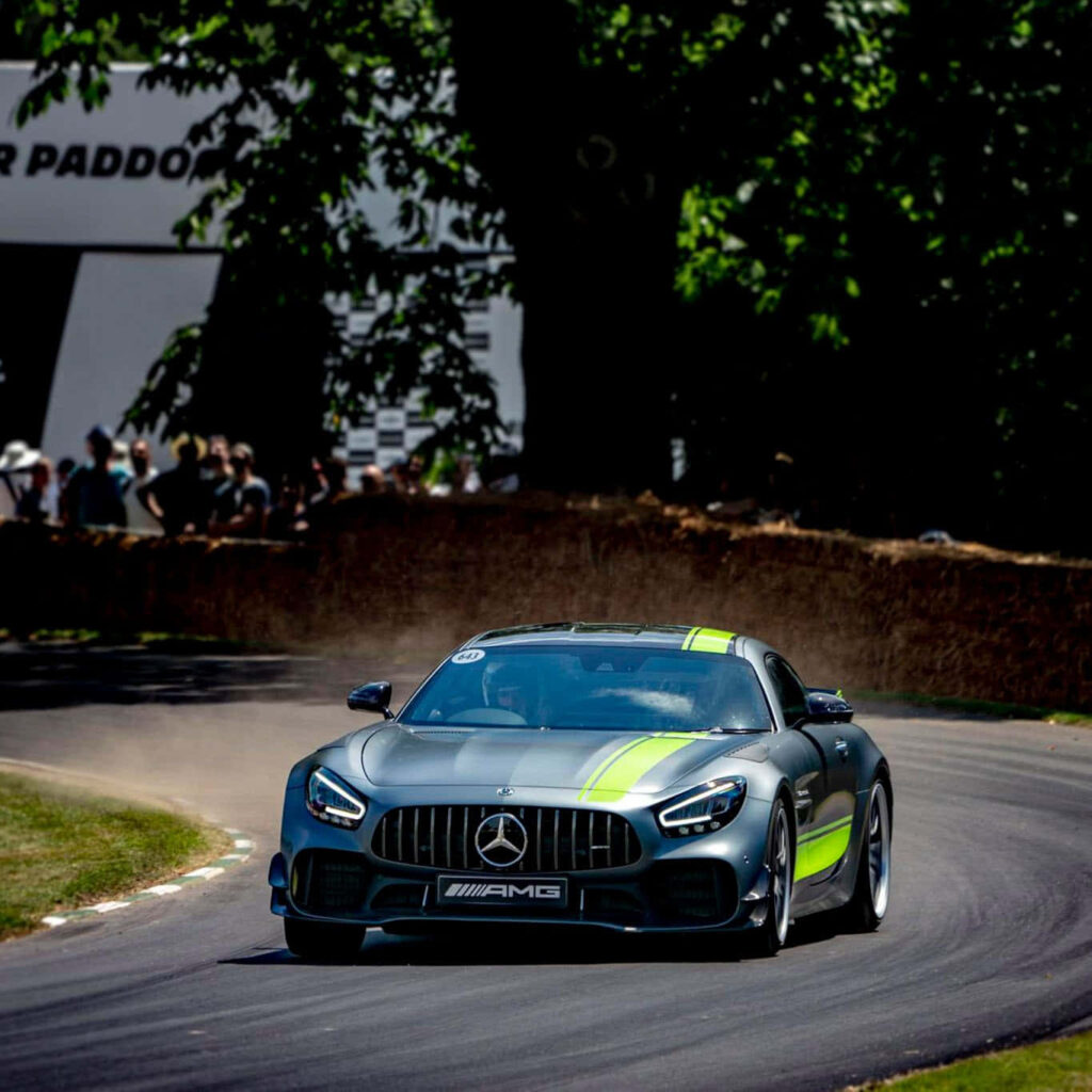 Mercedes AMG GT3 racing car with green stripes speeding on dusty racetrack near trees and crowd Wallpaper