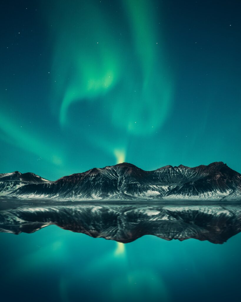 Vivid green northern lights swirl over snowy mountain peaks reflected in a calm lake at night Wallpaper