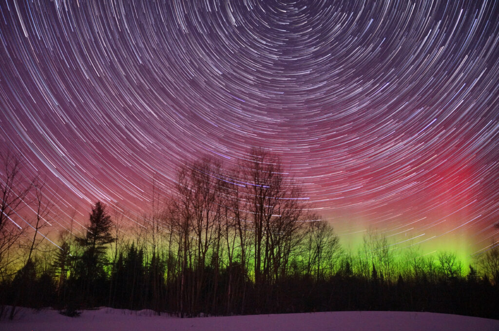 Silhouetted trees under vibrant northern lights and swirling star trails in colorful night sky wallpaper