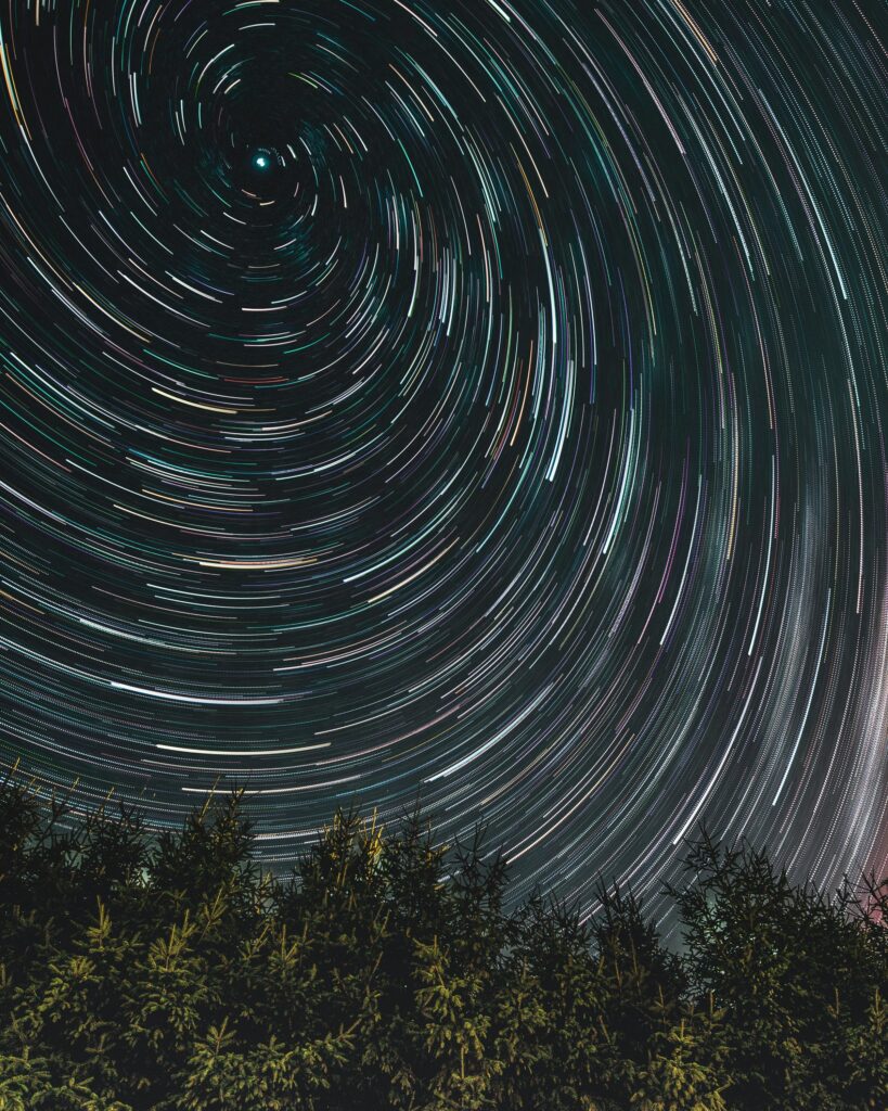 Star trail wallpaper with swirling light streaks above dark treetops in a serene night sky