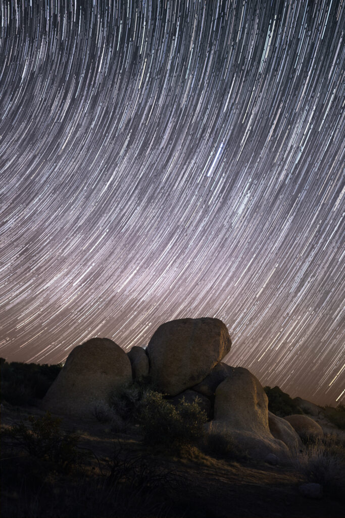 Night sky star trails swirl over large stones in serene earth tone landscape wallpaper background