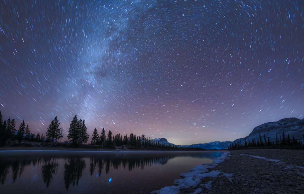 Starry night sky swirling above tranquil mountains pine trees and reflective calm lake wallpaper