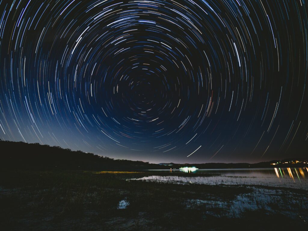 Starry night sky with swirling cosmic vortex over moonlit landscape reflected in calm water wallpaper