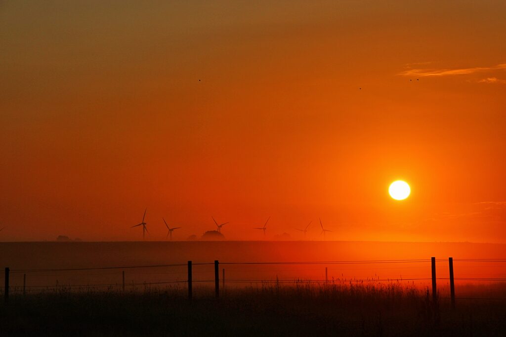 Sunset over open field with glowing orange sky and silhouettes of wind turbines and trees Wallpaper