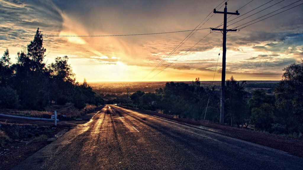 Golden sunset over hilltop road with wet pavement and vibrant cityscape under dramatic clouds Wallpaper