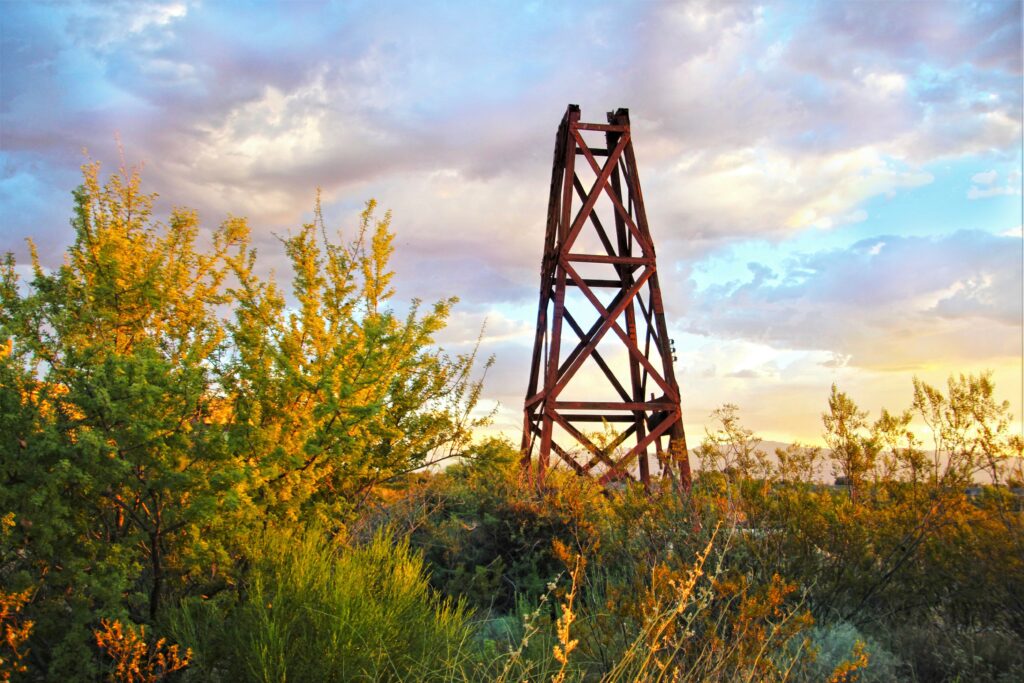 Metallic construction tower over dense green bushes under vibrant sky with dramatic clouds and sunlight Wallpaper