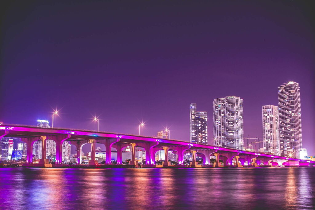 Miami skyline at night with MacArthur Causeway in purple pink hues over calm water reflection Wallpaper