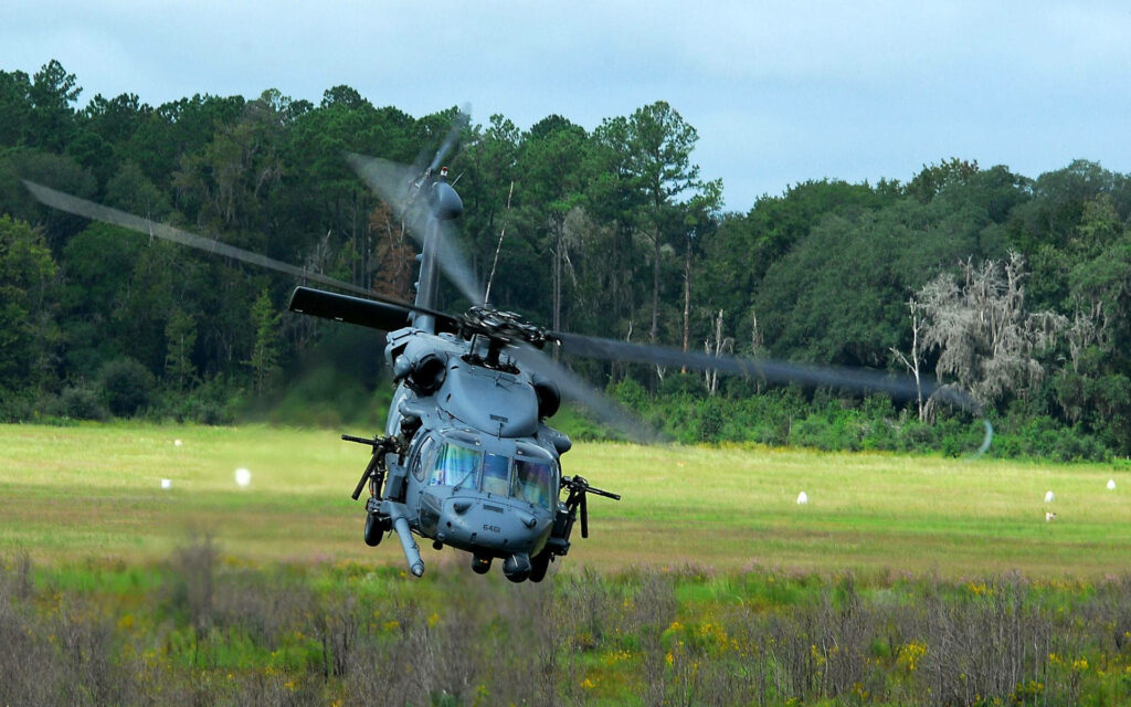 Military helicopter hovering low over grassy field with dense green forest and blue sky background Wallpaper