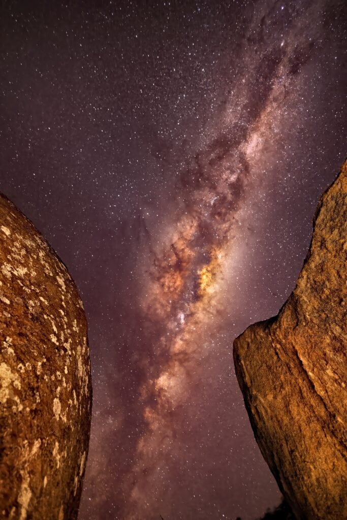 Milky Way galaxy over starry night sky with rugged rocks foreground and purple gold tones wallpaper