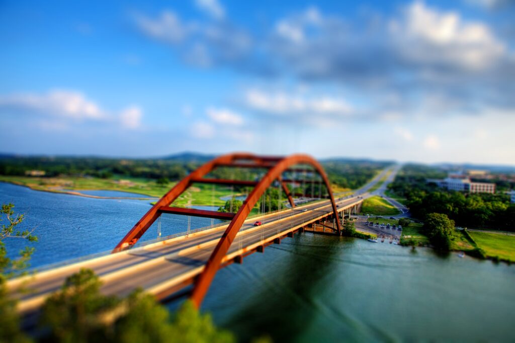 Tilt-shift wallpaper of red car on arched bridge over river with vibrant blue sky and green trees