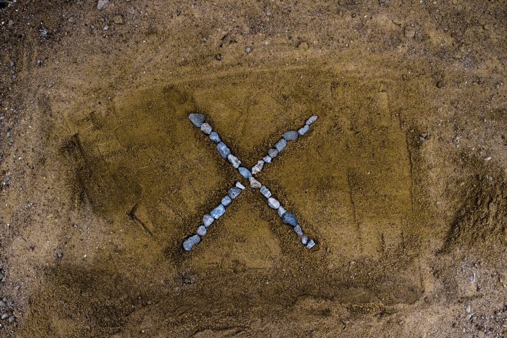 Bird's-eye view of letter X made from grey rocks on textured brown sandy background wallpaper