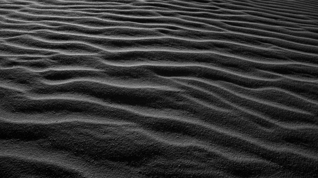Minimalist black and white wallpaper of rhythmic ripples on dark sand dunes with deep shadows