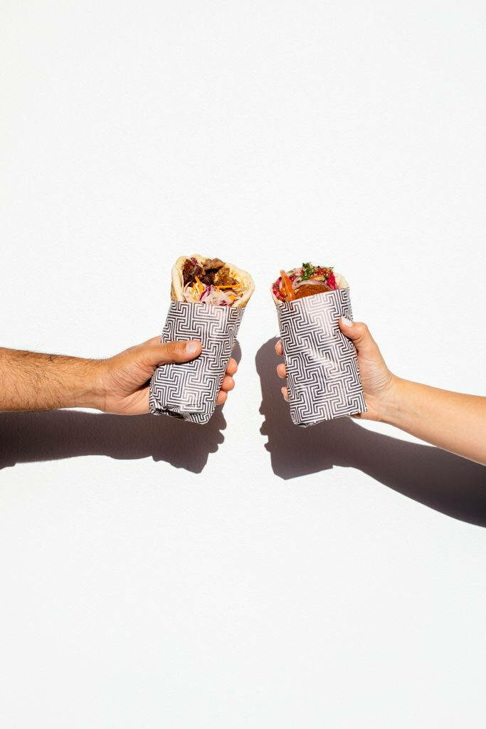 Two hands hold colorful burritos wrapped in black and white maze pattern paper on white background Wallpaper
