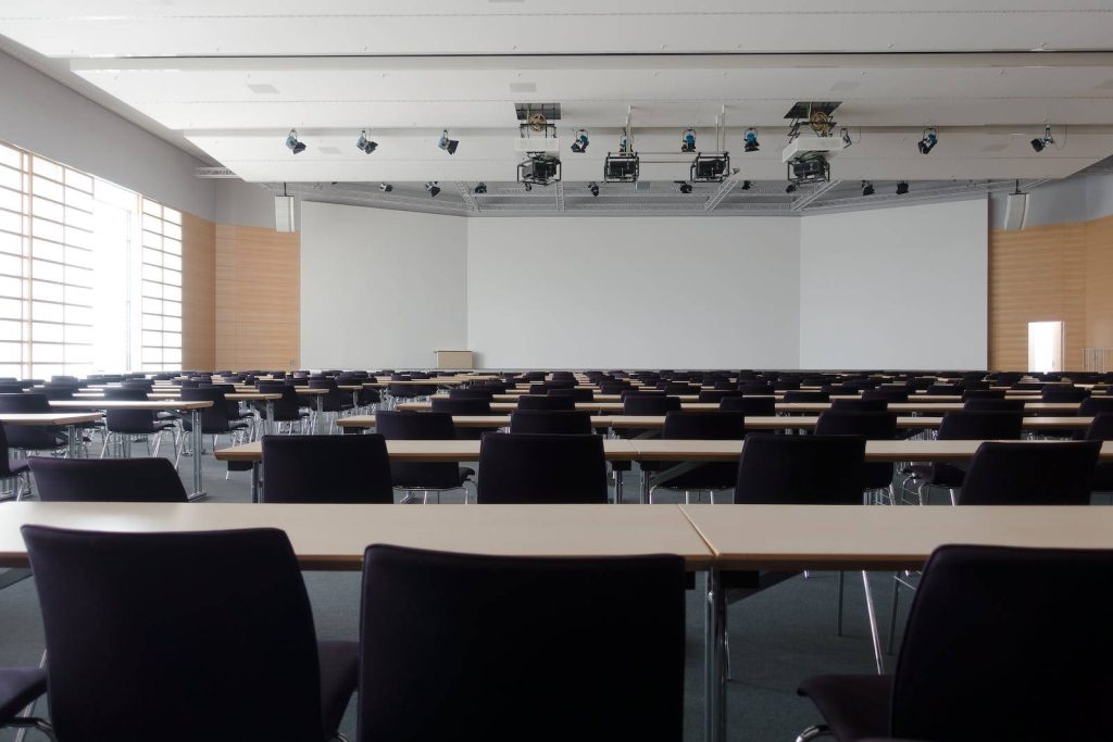 Bright modern lecture hall with black chairs wooden desks and large white projection screen Wallpaper