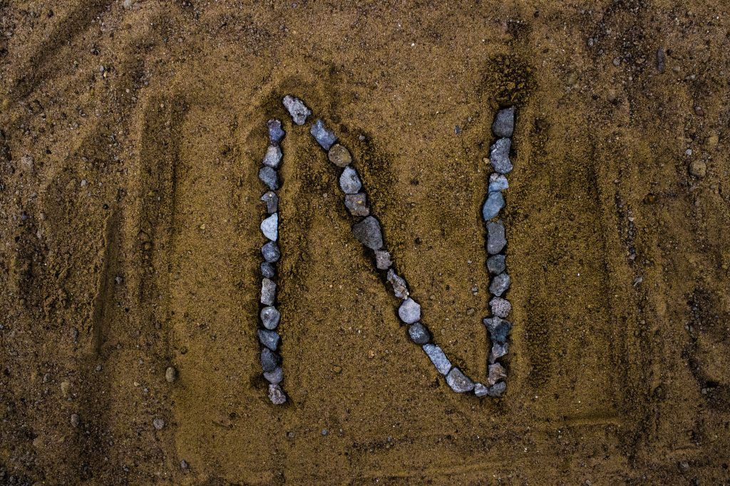Minimalist letter N made of gray pebbles on rich brown sand earthy textured wallpaper background