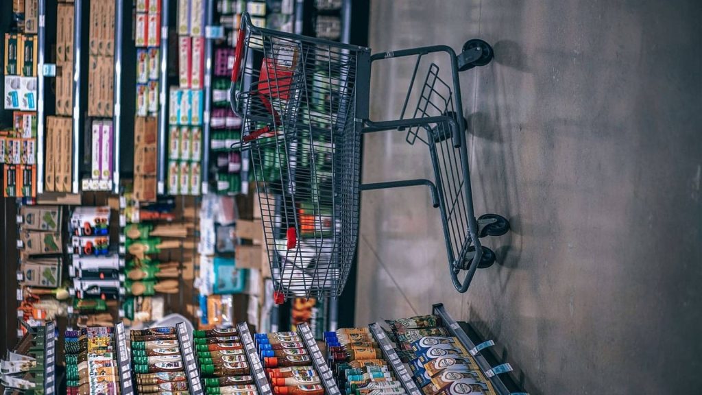Surreal metal shopping cart against grey grocery store wall with blurred supermarket shelves background Wallpaper