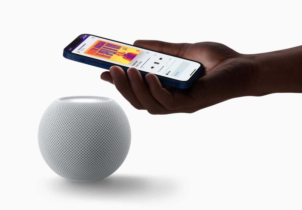 Hand holding iPhone with music controls above white Apple HomePod mini on bright studio background Wallpaper